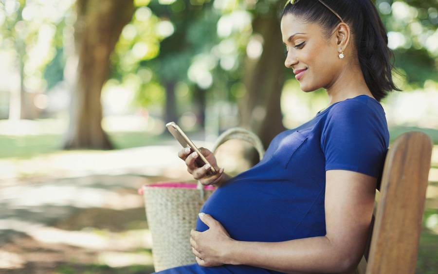A pregnant woman sits on a park bench, reading an article on her mobile phone.
