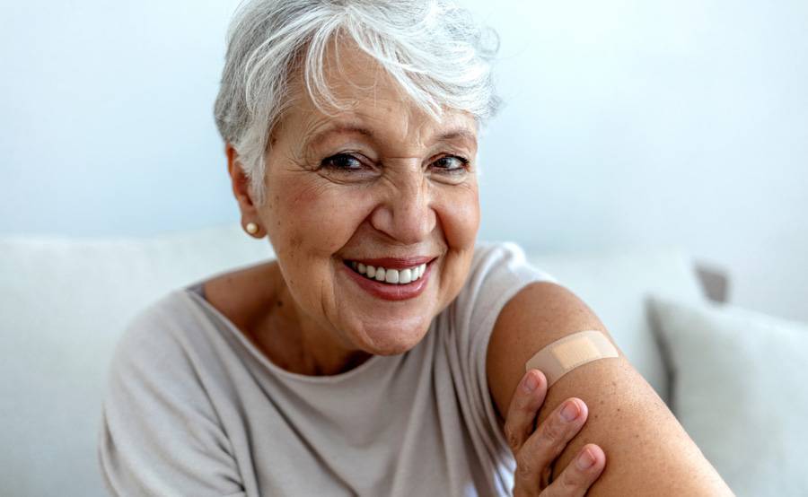 An older woman with gray hair smiles as she shows off her arm with a small bandage after her Shingles vaccine.