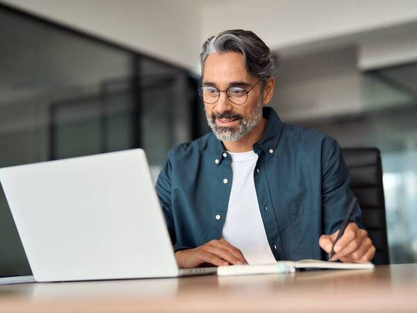 A middle aged man in glasses takes notes while looking at his laptop.